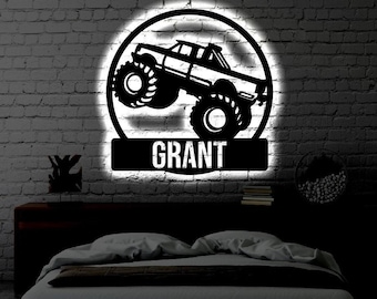 Puede incluir: Un letrero de pared circular de metal negro con la silueta de un camión monstruo con neumáticos grandes. Debajo del camión, una placa rectangular muestra el nombre GRANT en letras mayúsculas blancas. El letrero tiene una luz blanca de fondo.