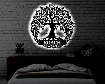 May include: A circular black metal wall art piece featuring a detailed tree of life design with intricate branches and leaves. A horizontal banner across the base displays the name ROACH in white capital letters, backlit by a bright glow.