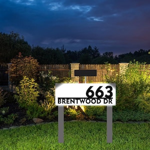 May include: A white sign with black text that reads "663 Brentwood Dr." The sign is mounted on two black metal posts and is illuminated by a light source.
