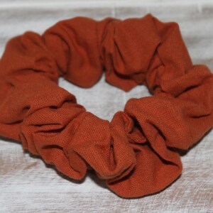May include: A burnt orange scrunchie made of soft fabric. The hair accessory is circular and gathered, with a textured appearance. This scrunchie is a stylish hair accessory for everyday wear.