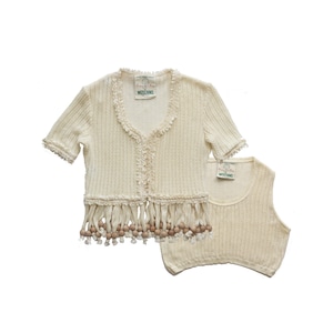 Puede incluir: Un cárdigan de crochet de color crema con ribete de flecos y cuentas de madera. El cárdigan tiene cuello en V y mangas cortas. La etiqueta dice "Moschino".