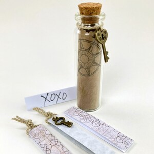 May include: A clear glass bottle with a cork stopper, filled with sand and a rolled paper featuring a compass rose design. A small bronze key charm hangs from the bottle. Three decorative bookmarks with ornate patterns and a small paper with the text "xoxo" are also present.