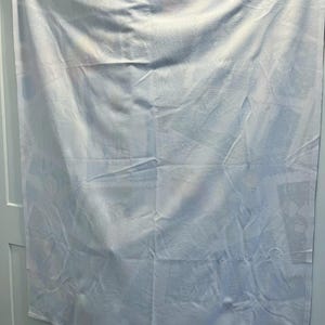 May include: White rectangular blanket with a subtle pattern, hanging on a white door. The fabric has soft folds and creases, suggesting a comfortable texture. The overall appearance is clean and simple, suitable for a bedroom.