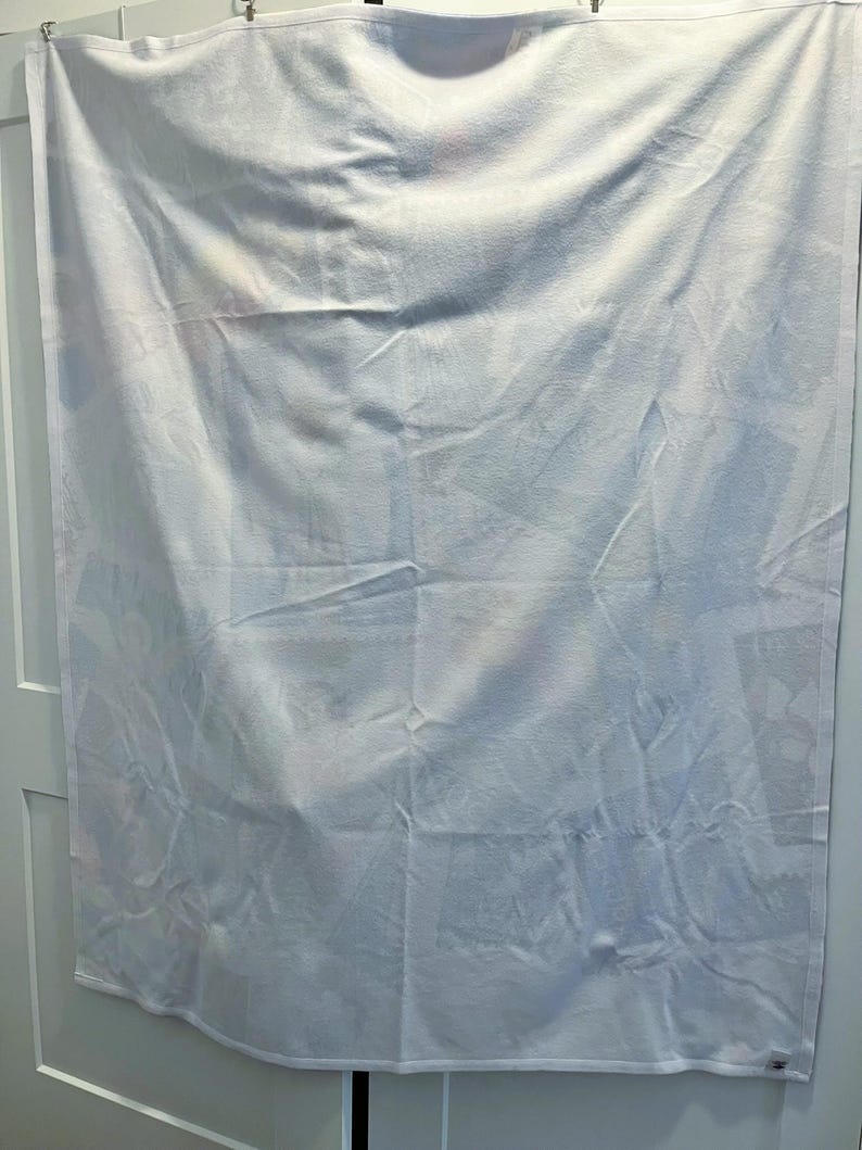 May include: White rectangular blanket with a subtle pattern, hanging on a white door. The fabric has soft folds and creases, suggesting a comfortable texture. The overall appearance is clean and simple, suitable for a bedroom.