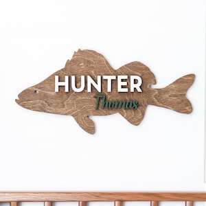 May include: A wooden fish-shaped wall decoration. It features the name "HUNTER" in white capital letters and the name "Thomas" in green script. The fish is a medium brown colour and is mounted on a white wall.