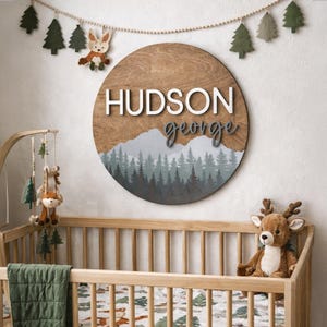 May include: A circular wooden sign with the name "HUDSON GEORGE" in white and grey lettering. The sign features a mountain and forest design in shades of grey and green. The sign is above a wooden cot with a stuffed deer.