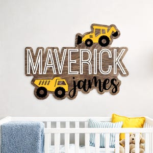 May include: Wooden sign with the name "MAVERICK" in white, with the name "james" in black script. Two yellow construction vehicles are featured. The sign is mounted above a white cot with a blue blanket and yellow cushion.