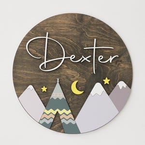 Peut inclure: Une pancarte ronde en bois avec le nom "Dexter" écrit en cursive blanche. La pancarte présente une scène de montagne fantaisiste avec un tipi, des étoiles et une lune croissante. Les montagnes sont peintes dans des tons de violet et de gris, et le tipi est décoré de rayures colorées.