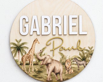 May include: A round wooden sign with a jungle theme. The sign features the name "Gabriel Paul" in white and gold lettering. The background features a giraffe, elephant, lion, and zebra.