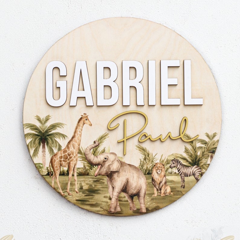 K&ouml;nnte beinhalten: Ein rundes Holzschild mit Dschungel-Motiv. Das Schild tr&auml;gt den Namen "Gabriel Paul" in wei&szlig;en und goldenen Buchstaben. Der Hintergrund zeigt eine Giraffe, einen Elefanten, einen L&ouml;wen und ein Zebra.