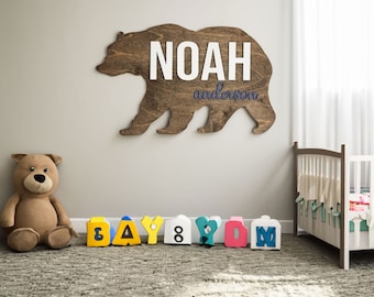 May include: Wooden bear cutout with the name "NOAH anderson" in white and purple lettering.  The bear is brown and is mounted on a white wall.  There are colorful wooden blocks that spell out "BABY" on the floor in front of the bear.