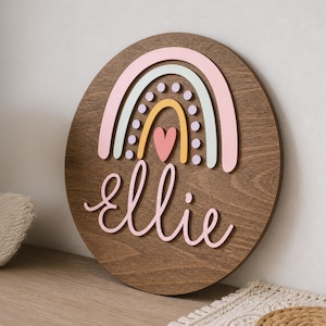 May include: A circular wooden sign with a rainbow design in pastel shades and the name "ellie" in cursive. The rainbow includes pink, green, and yellow arches with a pink heart. The sign is made of dark brown wood.