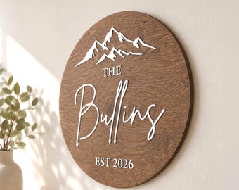 May include: A circular wooden sign with a dark brown wood grain finish featuring white raised lettering and graphics. The design includes a mountain range icon above the text "THE Bulling" and the date "EST 2026" printed in a white script font.