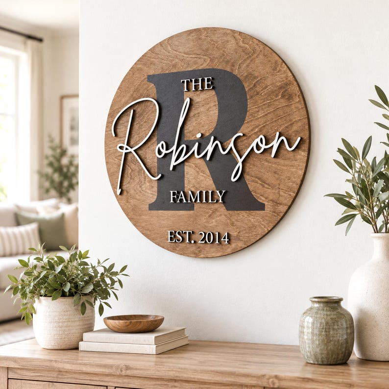 May include: A circular wooden wall sign featuring a large dark gray letter R, the white script text Robinson, and the white block text THE, FAMILY, and EST. 2014. The sign has a natural wood grain finish and is mounted on a white wall.