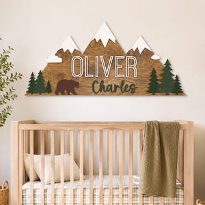 Op de afbeelding: Een houten wandbord in de vorm van een bergketen met witte, besneeuwde toppen, donkergroene dennenbomen en een silhouet van een bruine beer. Het bord bevat de naam OLIVER in witte blokletters en de naam Charles in donkergroene schrijfletters.