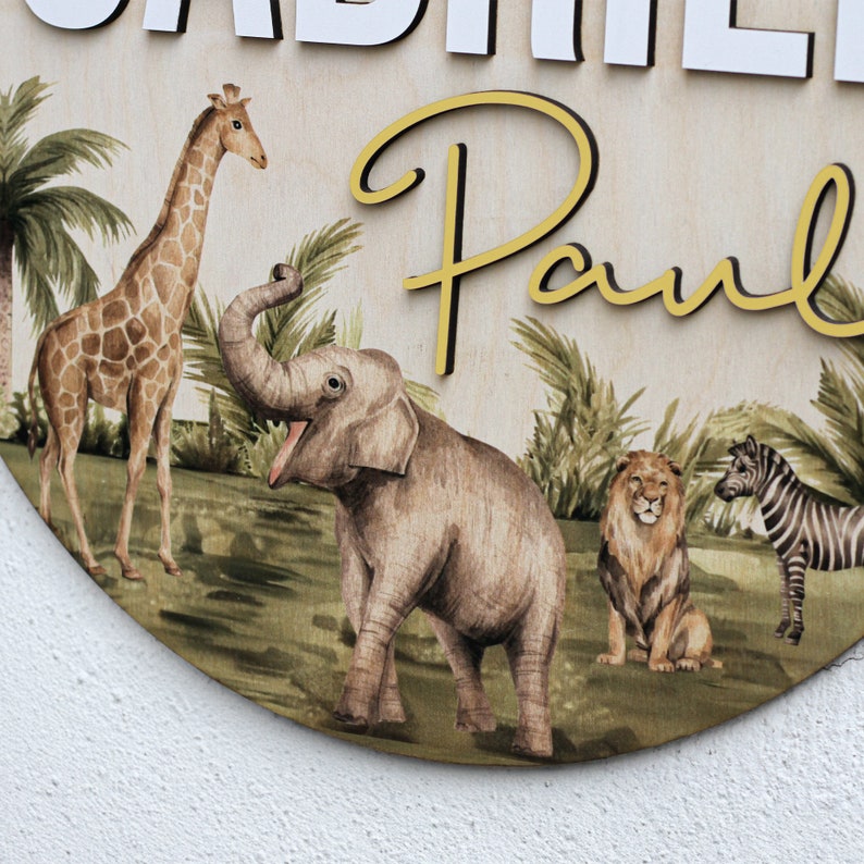 K&ouml;nnte beinhalten: Ein rundes Holzschild mit Dschungel-Motiv, auf dem eine Giraffe, ein Elefant, ein L&ouml;we und ein Zebra zu sehen sind. Das Schild tr&auml;gt die Namen "Gabriel" und "Paul" in goldener Schrift.
