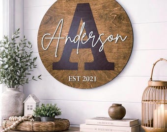 May include: A round wooden sign with a dark brown stain features the name "Anderson" in white script above the letter "A" in a dark brown color. The text "EST 2021" is below the name. The sign is displayed on a white wall.