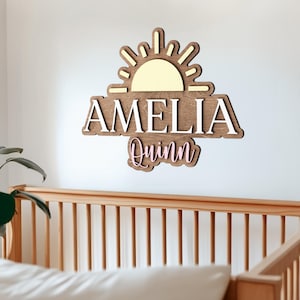May include: A wooden wall sign featuring a sun icon at the top with a pale yellow center. The name AMELIA is displayed in large white capital letters, and the name Quinn is written below in a pink script font, all mounted on a dark wood base.