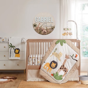 K&ouml;nnte beinhalten: Ein Babybett mit einem beige-braunen Bettw&auml;sche-Set im Safari-Stil. Die Bettw&auml;sche verf&uuml;gt &uuml;ber eine gesteppte Decke mit einer Giraffe, einem L&ouml;wen und einem Leoparden. Das Bett hat auch eine wei&szlig;e Wickelauflage mit einem Palmen- und L&ouml;wenmotiv. Das Bett ist mit einem runden Holzschild dekoriert, auf dem der Name "GABRIEL Paul" steht.