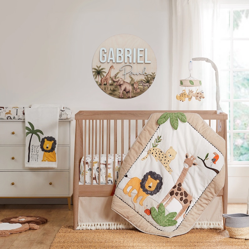 K&ouml;nnte beinhalten: Ein Babybett mit einem beige-braunen Bettw&auml;sche-Set im Safari-Stil. Die Bettw&auml;sche verf&uuml;gt &uuml;ber eine gesteppte Decke mit einer Giraffe, einem L&ouml;wen und einem Leoparden. Das Bett hat auch eine wei&szlig;e Wickelauflage mit einem Palmen- und L&ouml;wenmotiv. Das Bett ist mit einem runden Holzschild dekoriert, auf dem der Name "GABRIEL Paul" steht.