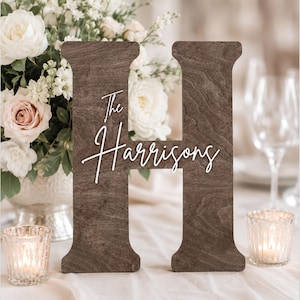 May include: A large, dark brown wooden letter H featuring the white script text The Harrisons centered on the front surface. The letter has a smooth, flat finish with visible wood grain patterns and a sturdy, block-like structure for upright display.