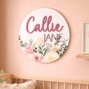 May include: A circular wooden sign featuring the name "Callie" in pink script above "Jane" in block letters. The sign is adorned with watercolour floral designs in pink, yellow, and green.
