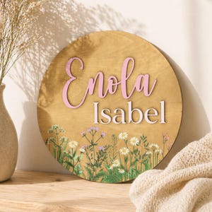 Op de afbeelding: Ronde houten plank met de naam "Enola Isabel" in roze letters. Het bord heeft een bloemenontwerp met madeliefjes en wilde bloemen. De achtergrond is een lichte houtnerf.
