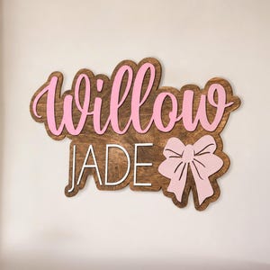 May include: Wooden sign with the names "Willow" and "Jade" in pink and white lettering. The sign has a brown wood grain background and a pink bow on the right side. A decorative item.