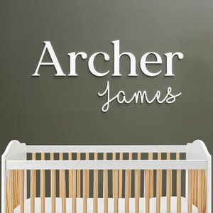 May include: White wooden name sign that reads "Archer James" in a nursery setting. The name is displayed above a white crib with wooden slats, set against a muted green wall. A decorative piece for a baby's room.