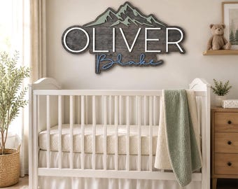 May include: A wooden mountain-shaped sign with the name "OLIVER" in white letters and "Blake" in blue script. The sign is mounted above a white crib with a green and cream blanket. A teddy bear sits on a shelf.
