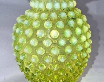 May include: A pale yellow hobnail vase with a ruffled rim. The vase has a round body covered in small, raised glass dots. The base is a solid, circular shape. The overall design is vintage and decorative.