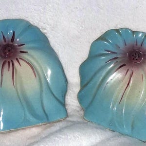 May include: A pair of ceramic salt and pepper shakers shaped like stylized flowers. Each shaker features a vibrant blue exterior that fades into a pale yellow center, accented by dark purple radiating lines and small holes at the top.