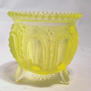 May include: A yellow glass bowl with three small legs. The bowl has a decorative beaded rim and ornate embossed patterns. The bowl is a light yellow color and appears to be made of glass.