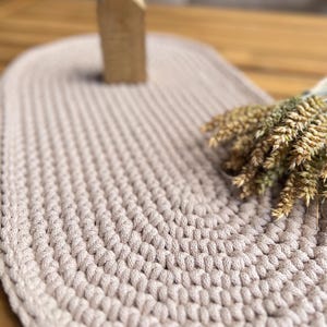 Könnte beinhalten: Eine ovale, hellbeige gehäkelte Matte mit einer dicken, strukturierten Kordelkonstruktion. Ein kleines, schlichtes, hausförmiges Holzornament steht aufrecht auf der Matte, und ein Bündel getrockneter Weizenähren liegt auf der rechten Seite.
