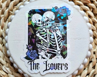 May include: A holographic sticker featuring a skeletal couple embracing, with the text "The Lovers" below. The background is a glittery, iridescent design with blue roses.