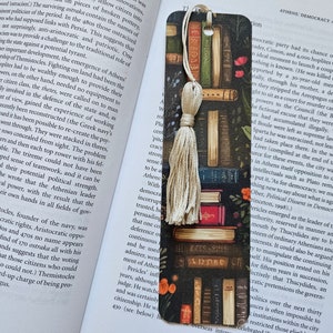 May include: A bookmark with a stack of colorful books, a tassel, and floral accents. The bookmark is designed to look like a bookshelf.