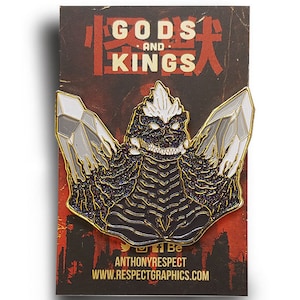 May include: A black and gold enamel pin featuring a Godzilla-like monster with white spikes on its head and two large crystals behind it. The pin is on a card with the text "Gods and Kings" in red and black lettering.