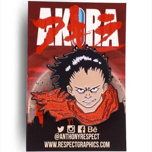 Peut inclure: Une épingle en émail rouge et noire représentant un personnage de l'anime Akira. Le personnage a les cheveux noirs, une écharpe rouge et une expression sérieuse. L'épingle est sur un fond rouge avec le mot "Akira" en lettres blanches.