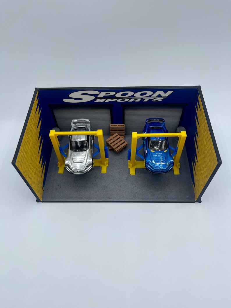May include: A miniature garage diorama with a blue and yellow exterior. The garage has two car lifts with a silver and a blue car on them. The garage has the text "SPOON SPORTS" on the front.