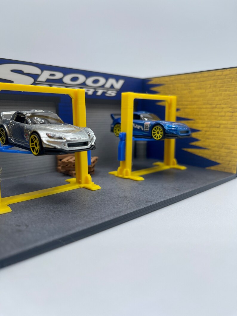 May include: A miniature model garage with two yellow lift platforms. A silver Honda S2000 and a blue Subaru Impreza are on the lifts. The garage has a blue and yellow wall with the text "SPOON PARTS" on it.