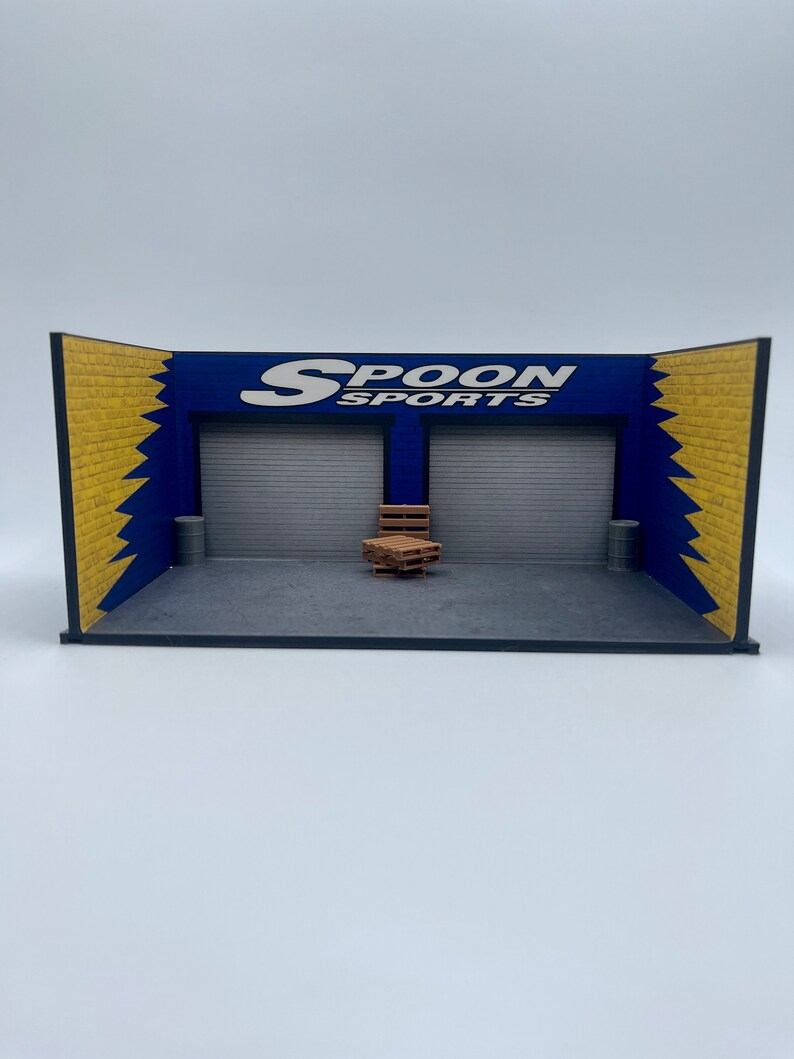 May include: A miniature diorama of a garage with a blue and yellow brick wall, a roll-up door, and the words "SPOON SPORTS" in white lettering. There are wooden pallets stacked in the center of the garage.