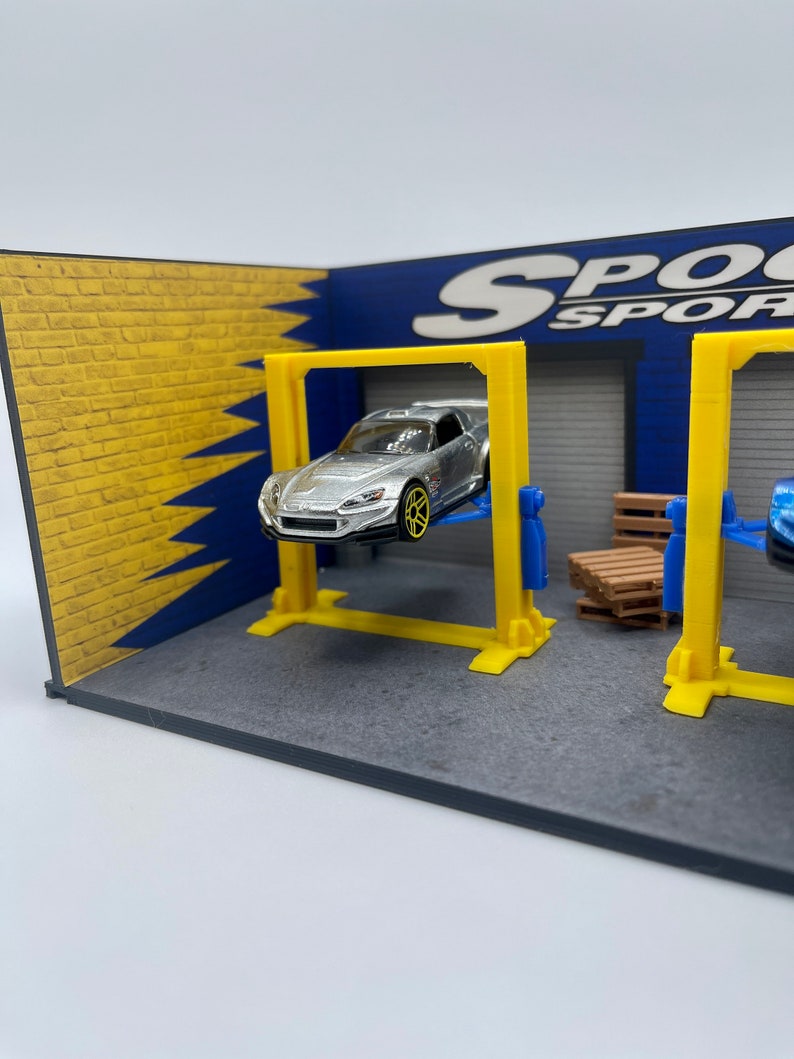 May include: A silver toy car is lifted on a yellow and blue car lift in a miniature garage setting. The garage has a brick wall and a blue wall with the text "SPOOK SPORT" on it.