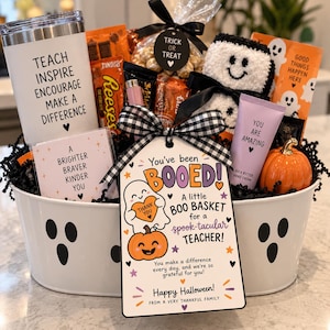 May include: A white oval metal basket with ghost faces containing a white tumbler with text, a bag of treats with a Trick or Treat tag, a plush ghost, a tube of hand cream, a small orange pumpkin, and a card reading You've been BOOED!