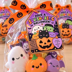 May include: A clear plastic treat bag tied with an orange ribbon featuring black jack-o'-lanterns. The bag contains a white ghost, an orange pumpkin, and a purple bat figurine, plus a tag reading "HAVE A SQUISH-TASTIC HALLOWEEN!" with cute characters.