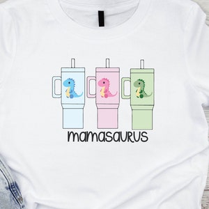 May include: White t-shirt with a graphic of three colorful cups with dinosaurs holding straws. The text "mamasaurus" is printed below the cups.