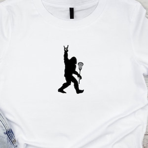 May include: White t-shirt with a black silhouette of a Bigfoot holding a lacrosse stick and making a rock and roll hand gesture.