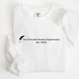 Op de afbeelding: Witte longsleeve sweatshirt met zwarte tekst die "The Tortured Nurses Department. est. 2024" luidt en een zwarte veerpen-afbeelding.