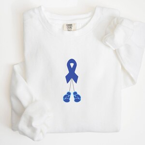 May include: White long-sleeved sweatshirt with a blue ribbon and blue boxing gloves printed on the front.