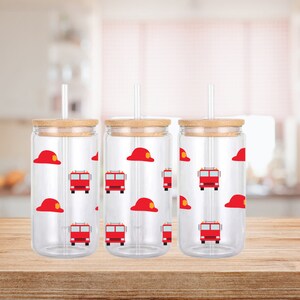 Puede incluir: Tres vasos de vidrio transparente con tapas de bambú y pajitas. Cada vaso está decorado con un patrón de camiones de bomberos rojos y cascos de bombero. Los vasos están sobre una superficie de madera.