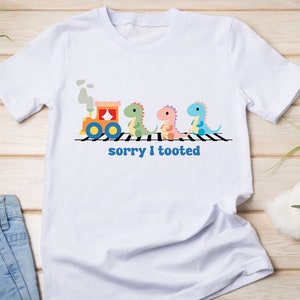 May include: White t-shirt with a cartoon train and dinosaurs on a railroad crossing. The text "sorry I tooted" is printed below the image.